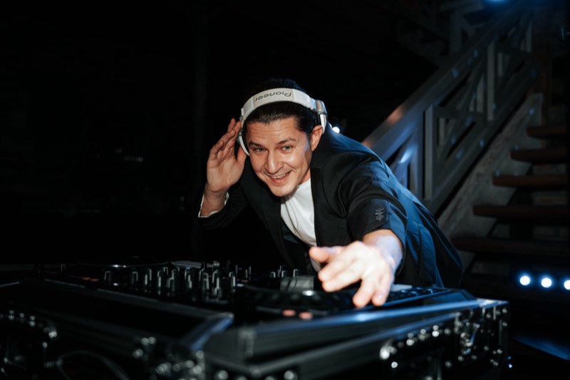 DJ Edouard lacht in die Kamera und zeigt auf sie, eine Hand am Kopfhörer