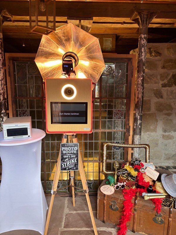 Frontalansicht der goldenen E-EVENTS Fotobox mit leuchtendem Ringlicht und Touchscreen „Zum Starten berühren“, darüber der goldene Blitzschirm, davor die Kreidetafel und der Requisitenkoffer mit Federboas und Schildern