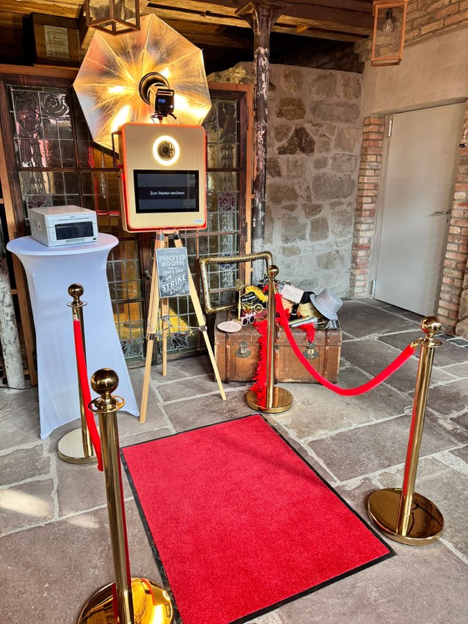 Goldene E-EVENTS Fotobox mit Ringlicht und goldenem Blitzschirm, daneben Sofortdrucker, Requisitenkoffer, roter Teppich und goldene Absperrpfosten mit roten Kordeln