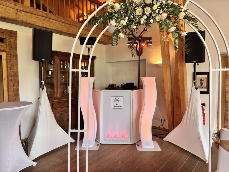 Weißes DJ-Pult mit Lautsprechern und Blumenbogen, aufgebaut für eine Trauung in einem Holzsaal